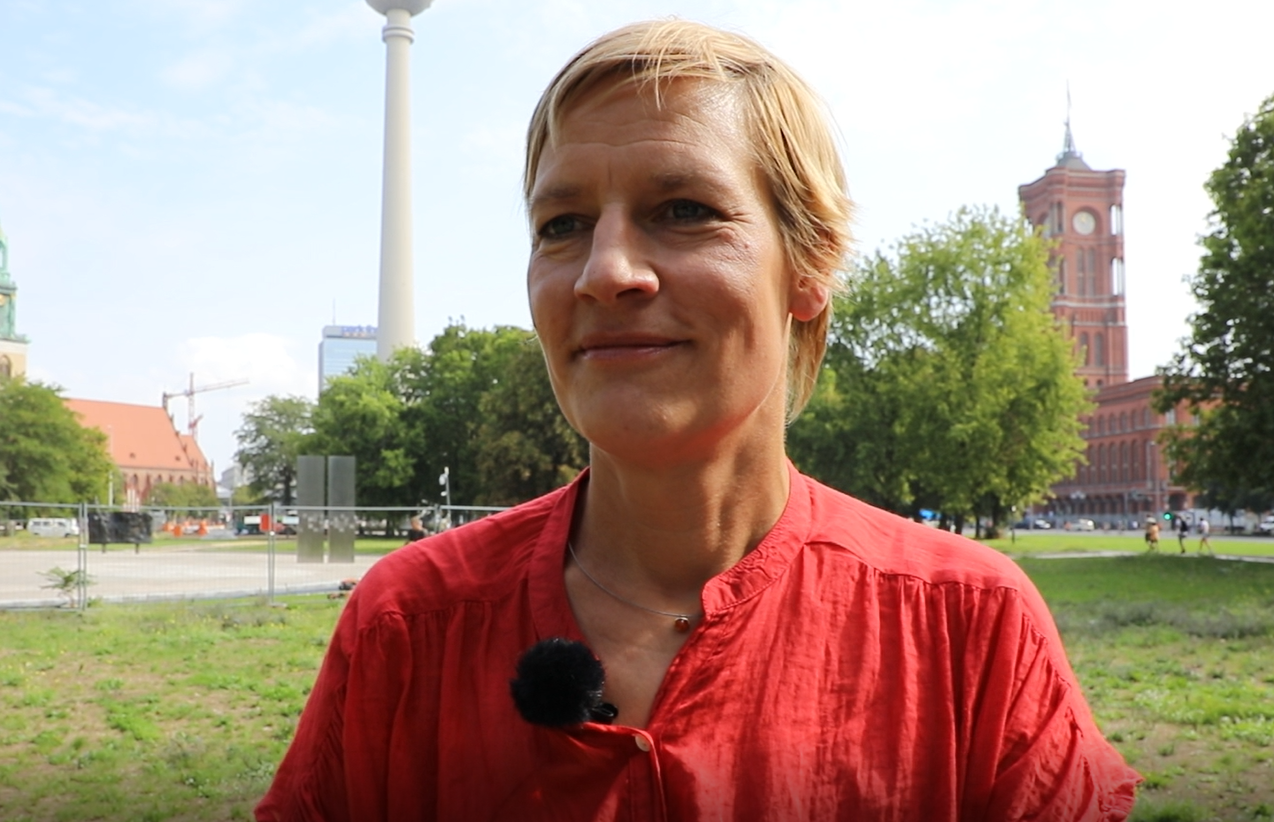 Die Mitarbeiterin im Bereich Projekte Freiraum/Infrastruktur steht vor dem Fernsehturm und dem Roten Rathaus in Berlin und trägt ein rotes Hemd