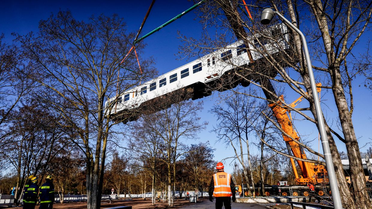Ein ausrangierter weißer Bahnwaggon wird von einem Kran über Bäume hinweg gehoben.