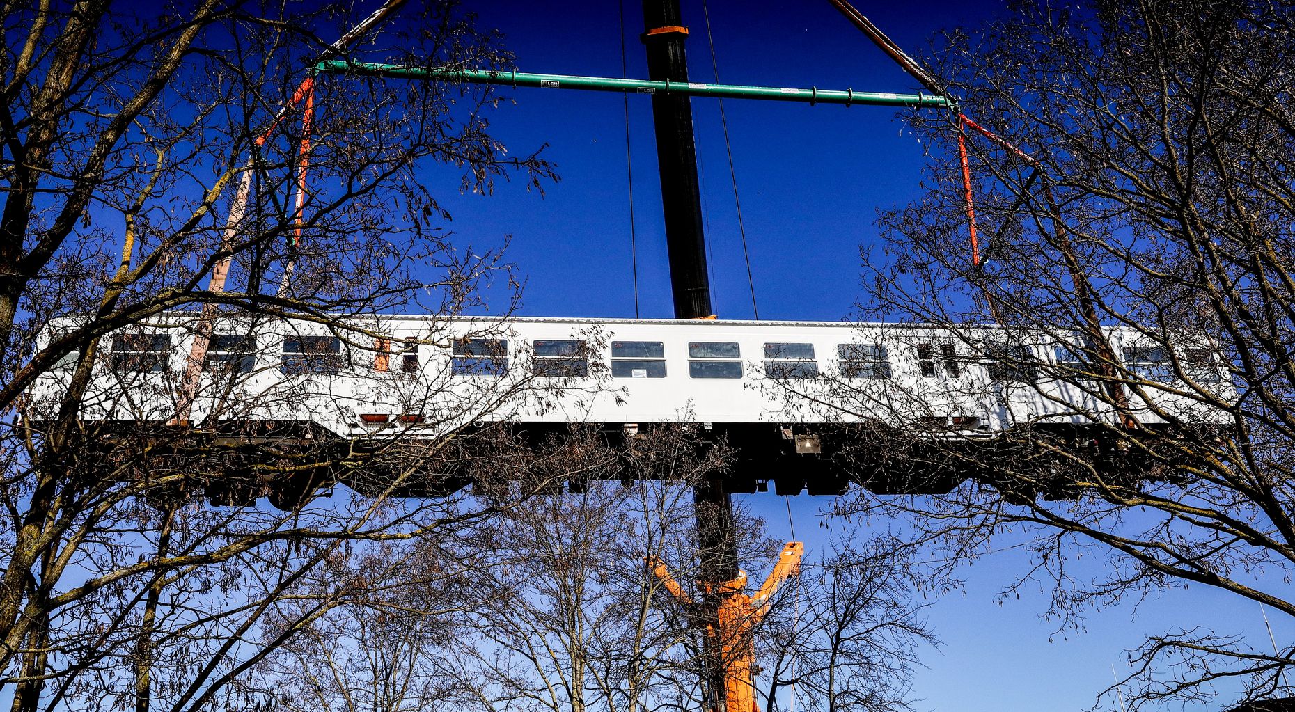 Ein ausrangierter weißer Bahnwaggon wird von einem Kran über Bäume hinweg gehoben.