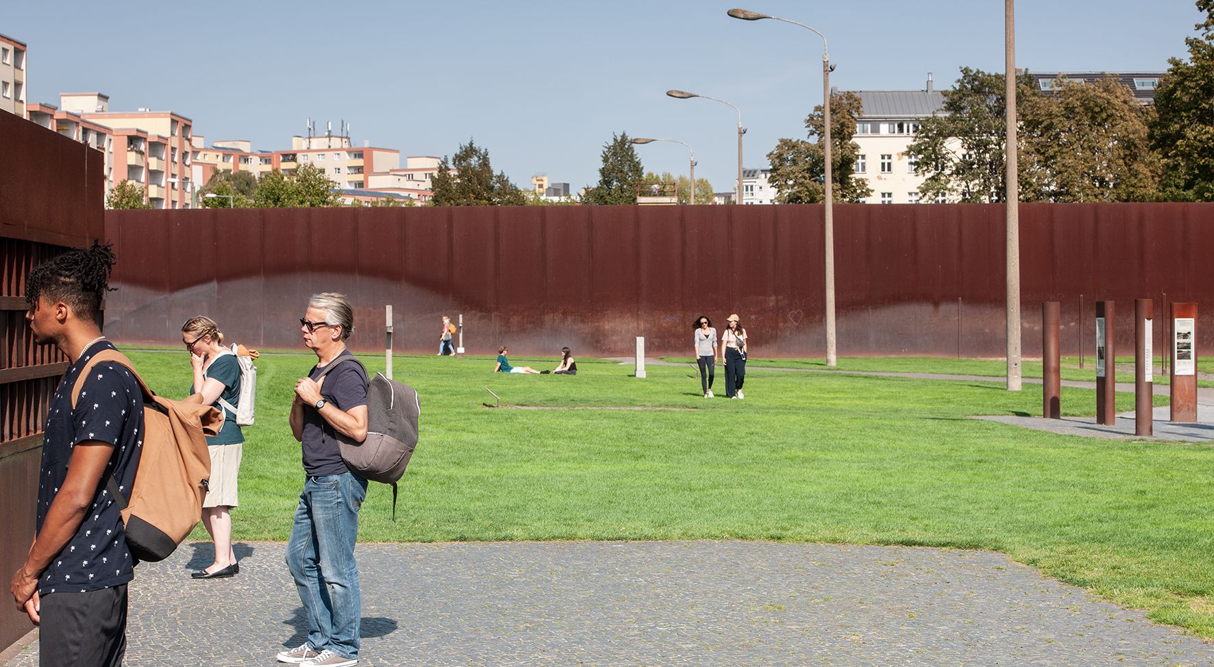Das Bild zeigt eine Wiese umgeben von einer rostbraunen Mauer. Auf der Wiese sitzen Menschen, spazieren darüber. Andere Menschen schauen auf die Mauer.