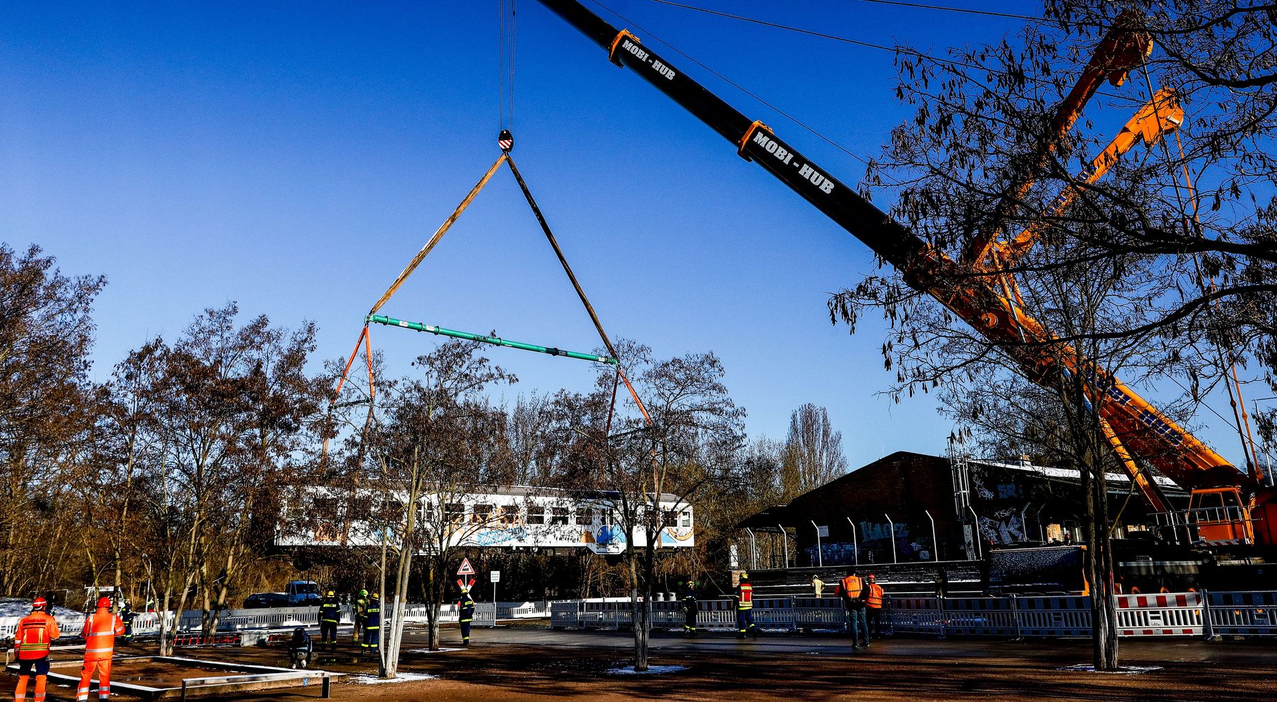 Ein ausrangierter weißer Bahnwaggon wird von einem Kran über Bäume hinweg gehoben.