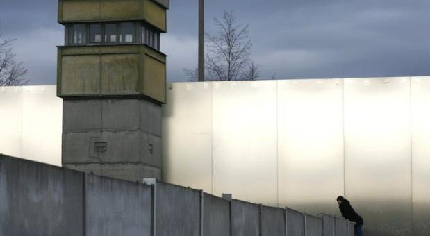 Das Bild zeigt eine Mauer mit Wachturm. An der Mauer stützt sich ein Mensch hoch.