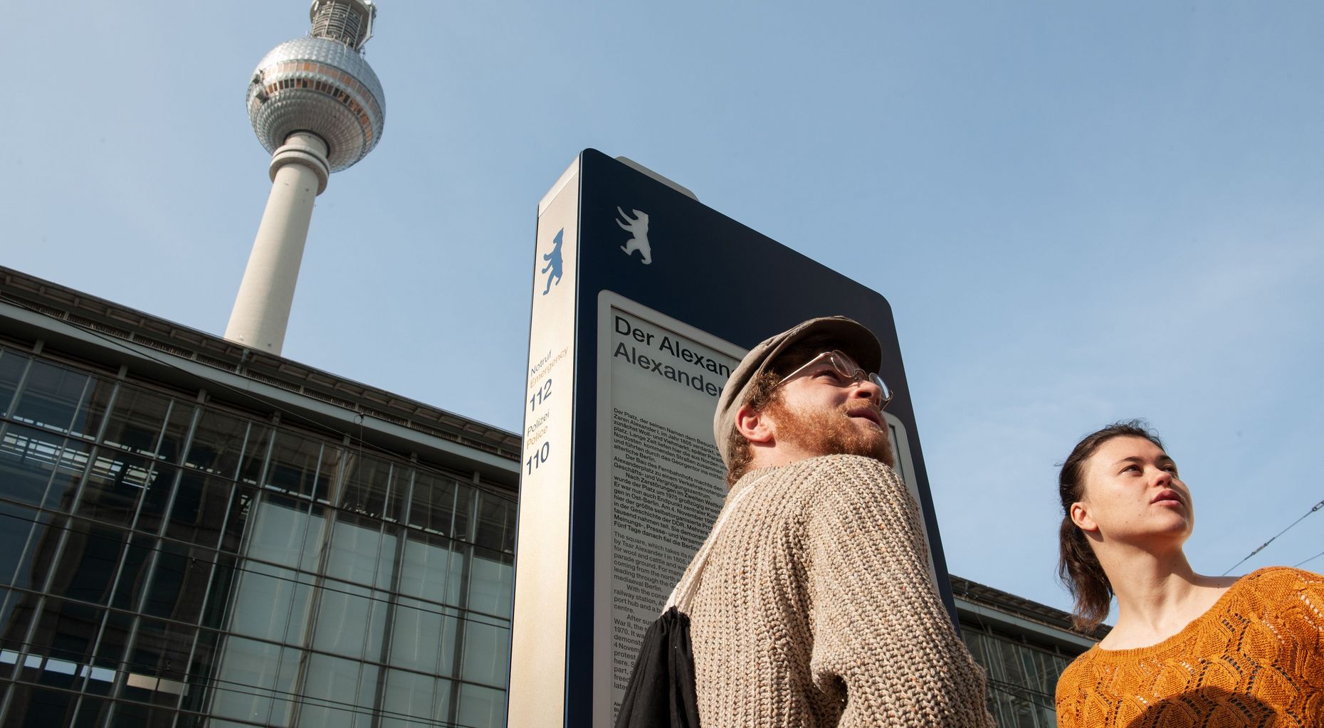 Ein Mann und eineFrau stehen vor der saphirblauen Informationsstele am Alexanderplatz.